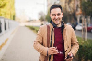 Portrait of a handsome businessman smiling while holding eyeglasses and phone in his hands and standing on the street.